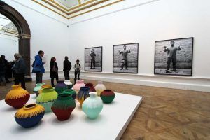 Ai Weiwei, Coloured Vases, 2015, Zwölf Vasen aus der Han Dynastie (206 vor Chr.- 220 nach Chr.) und vier neolithische (5000-3000 vor Chr.) mit Industriefarbe bemalt, verschiedene Größen, Privatsammlung und Sammlung von Lisa und Danny Goldberg. Im Hintergrund: Dropping a Han Dynasty Urn, 1995, Triptychon, Schwarz-weiß Fotografien, je 199,9 x 180 cm, Ausstellungsansicht Royal Academy, London 2015, Foto: Alexandra Matzner.